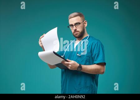 Waist up portrait of mature doctor holding clupboard while posing against blue background in studio, copy space Stock Photo