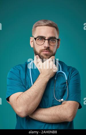 Vertical waist up studio portrait shot of pensive young adult doctor wearing blue uniform and eyeglasses touching his chin thinking of something