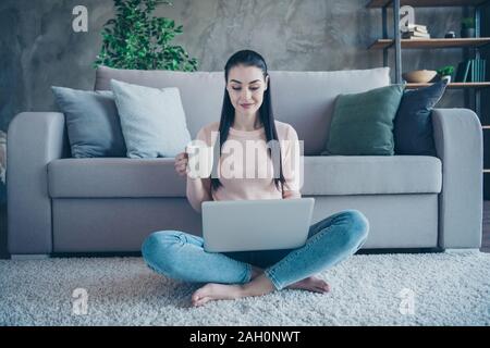 Photo of pretty lady with hot beverage holding notebook on knees leaning sofa sitting floor on comfy fluffy carpet wearing pullover and jeans in Stock Photo