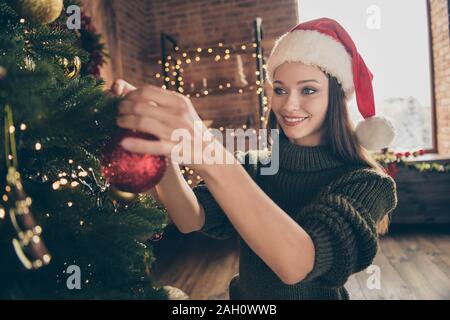 Close up photo of positive cheerful girl in santa claus cap knitted sweater enjoy christmas celebration hang red big bauble on evergreen tree in house Stock Photo
