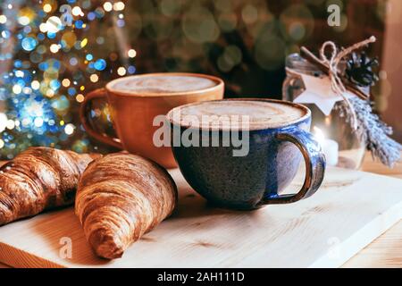 Perfect breakfast of croissants and cappuccino in a cafe. Blue and yellow ceramic cups of cappuccino and croissants on wooden table with Christmas lig Stock Photo