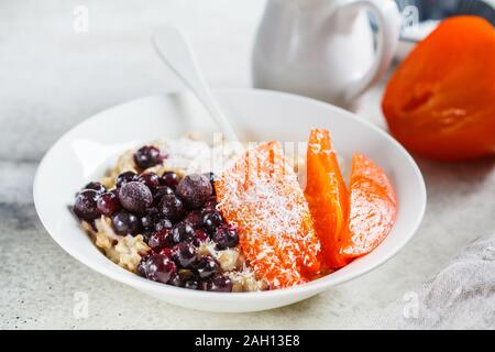 Oatmeal with persimmons, coconut and berries in a white plate on a gray background.
