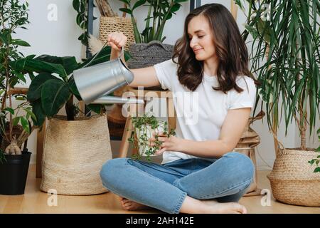 Young brunette sitting on a floor, watering potted plants in her home greenhouse Stock Photo