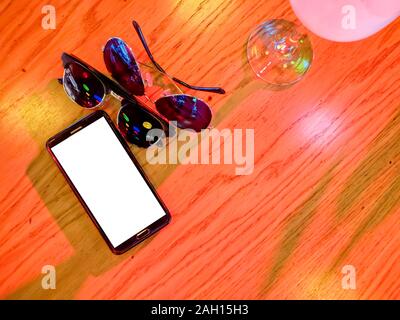 Phone and a couple glasses. Modern black smart phone with white isolated display for mockup in wooden table. Cocktail, glasses. Stock Photo