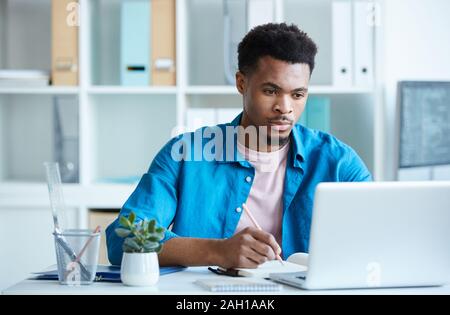 Serious African young man in casual clothing sitting at the table looking at monitor of laptop and making notes at office Stock Photo