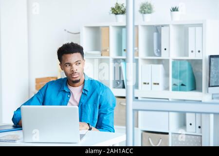 African pensive man in casual clothing sitting at the table in front of laptop and thinking about something at office Stock Photo