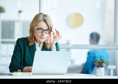 Serious mature female leader in eyeglasses sitting at the desk and working online she typing on laptop computer at office Stock Photo