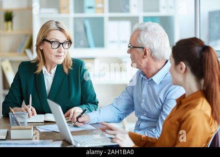 Mature businesswoman in eyeglasses talking to her colleagues and they discussing business plan at the table at business meeting Stock Photo
