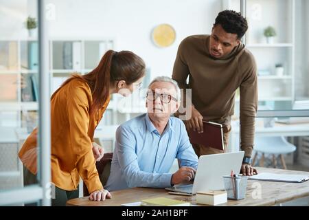 Young business people talking to their mature experienced colleague while he sitting at the table and using laptop Stock Photo