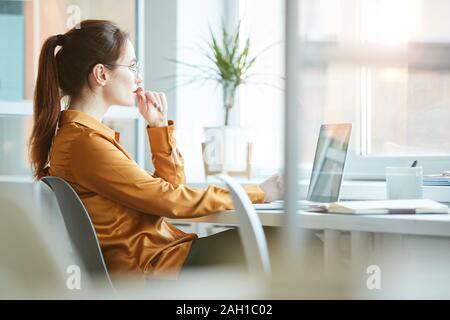 Young woman sitting at her workplace in front of the window with pensive sight and thinking about something while working on laptop Stock Photo