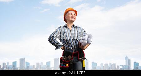 Attractive female engineer in hardhat standing with technical blueprints. Portrait of happy young architect in checkered blue shirt on background of c Stock Photo