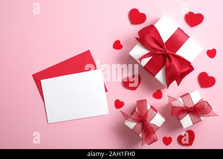 Love Letter with blank paper note and white gift boxes with red ribbon bow on pastel pink background decorated red hearts. Flat lay, top view. Saint V Stock Photo