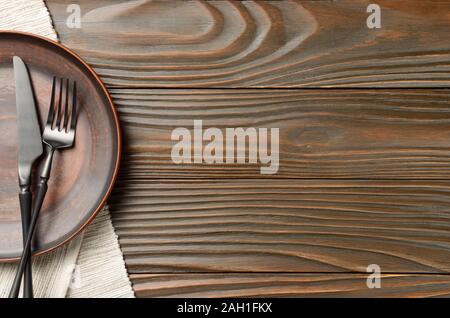 Top view at empty clay plate knife and fork aside on grey napkin on brown wooden kitchen table Stock Photo