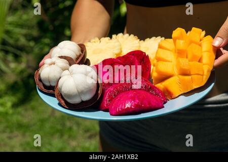 A girl in the green jungle is holding a plate with freshly cut and peeled tropical fruits. Stock Photo