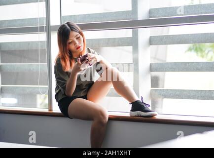 Young highschool girl sitting by the window using smartphone. Bored student text friends in the class. Stock Photo