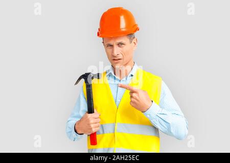 Construction. Mature man in hardhat and uniform standing isolated on white pointing at hammer concerned Stock Photo