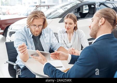 Car Salon. Young couple sitting at table with agent presenting car on digital tablet while man and woman looking at screen concentrated Stock Photo