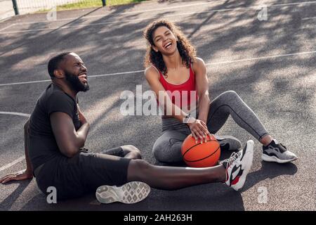 Outdoors Activity. African couple sitting with ball on basketball court laughing cheerful having fun Stock Photo
