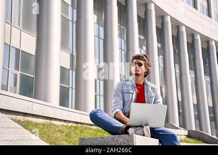 Freestyle. Guy in jean jaket and sunglasses outdoors sitting free time working freelance on laptop thinking of new creative idea Stock Photo