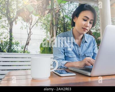 Beautiful happy asian woman short hair wearing blue jeans shirt using laptop computer and her smartphone put on wooden table in cafe with green backgr Stock Photo