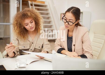 Horizontal medium eye level shot of two attractive ethnically diverse young women working together on project in modern office Stock Photo