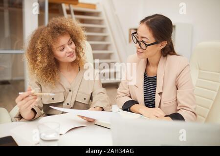 Horizontal eye level shot of two beautiful ethnically diverse young women working together sitting at office table Stock Photo