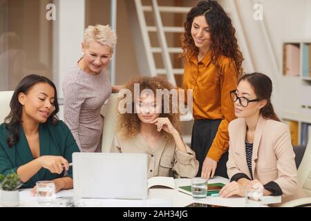 Horizontal portrait of five charming female colleagues watching something on laptop while working on business project together Stock Photo