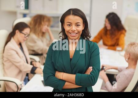 Charming African American woman standing with arms closed looking at camera smiling with colleagues coworking behind her, waist up portrait Stock Photo