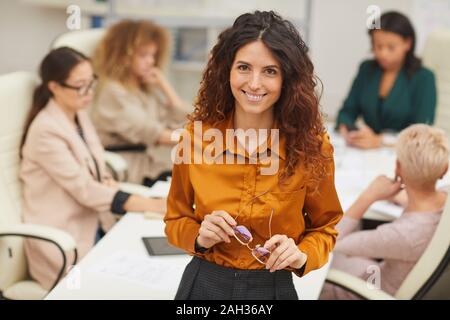 Charming Caucasian woman standing with with eyeglasses taken off looking at camera smiling with colleagues coworking behind her medium portrait Stock Photo