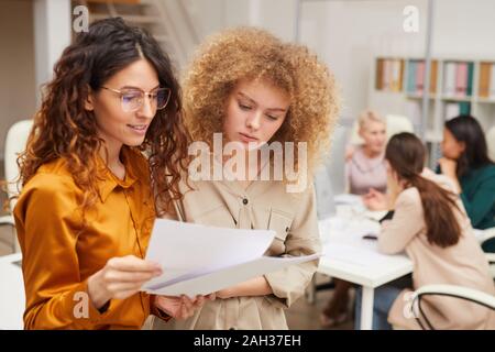 Two young businesswomen looking at documents, their colleagues coworking behind them Stock Photo