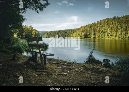 Bench in front of a lake with clouds on the sky and trees behind in the bavarian forest Stock Photo