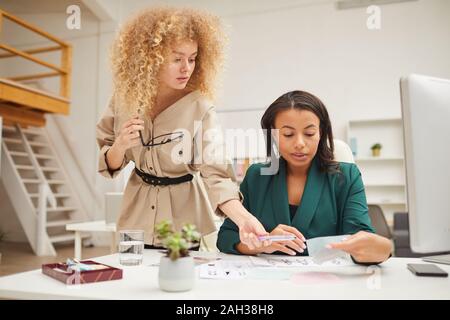 Two ethnically diverse young women coworking on new fashion collection fabric choice in modern office Stock Photo