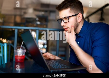 Thoughtful young attractive hipster freelancer working remotely on laptop in loft cafe. Businessman browsing internet at coffeshop. Blogger posts to s Stock Photo