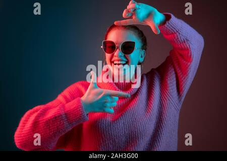 Framing. Caucasian woman's portrait isolated on gradient studio background in neon light. Beautiful female model with sunglasses, red hair. Concept of human emotions, facial expression, ad. Stock Photo