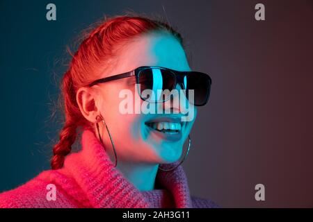 Close up, smiling. Caucasian woman's portrait isolated on gradient studio background in neon light. Beautiful female model with sunglasses, red hair. Concept of human emotions, facial expression, ad. Stock Photo