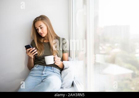 Beautiful woman drinking coffee use mobile phone and looking through window while sitting at windowsill at home Stock Photo