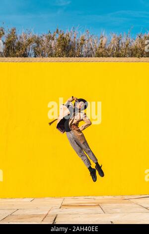 Young man dancing in front of yellow wall, jumping mid air Stock Photo