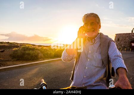 Senior man using smartphone on bicycle at sunset, Tenerife Stock Photo
