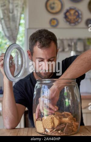 Man sitting in kitchen, looking in cookie jar, taking cokkie Stock Photo