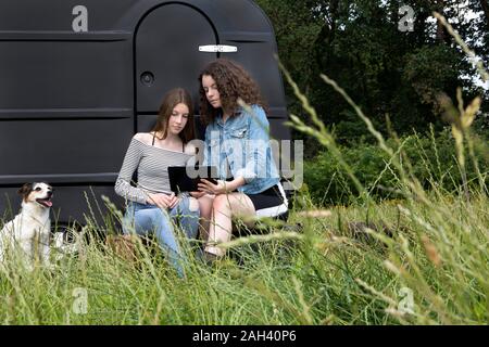 Two friends sitting on a meadow in front of black caravan looking at digital tablet Stock Photo