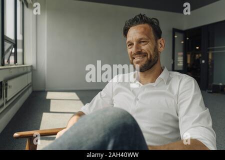 Smiling mature businessman sitting in armchair in empty office Stock Photo