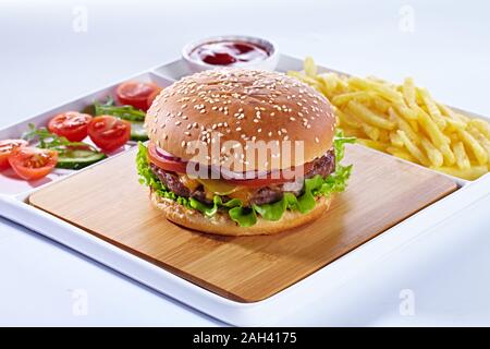 Juicy tasty hamburger on a wooden cutting board with french fried fries, vegetables and ketchup. Isolated composition on a white background and white Stock Photo