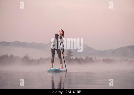 Woman stand up paddling on lake Kirchsee at morning mist, Bad Toelz, Bavaria, Germany Stock Photo
