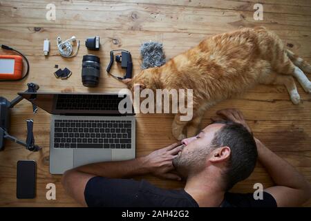 Exhausted man sleeping on table with ginger cat, laptop and photografic equipment Stock Photo