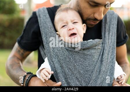 Young father carrying his crying baby son in a baby sling Stock Photo