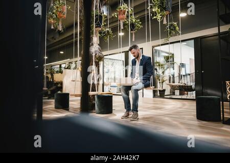 Businessman using laptop in coworking space, sitting on swing Stock Photo