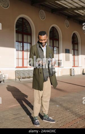 Stylish man with reusable cup using smartphone while waiting for the train Stock Photo