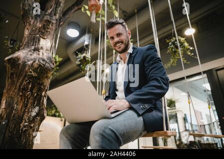 Businessman using laptop in coworking space, sitting on swing Stock Photo