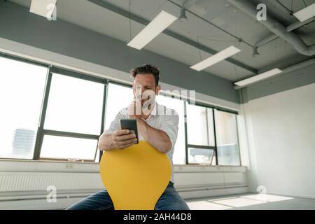 Mature businessman sitting on yellow chair in empty office using smartphone Stock Photo
