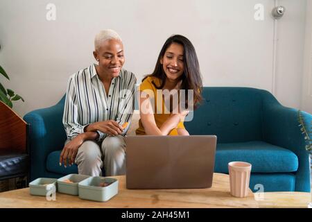 Two happy women sitting on couch using laptop together Stock Photo